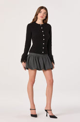 A woman is wearing a black, long-sleeved, button-up cardigan with a textured, fuzzy fabric and feathered cuffs. She is also wearing a high-waisted, dark gray, voluminous mini skirt with a gathered, bubble style. She has on black high-heeled shoes with ankle straps.
